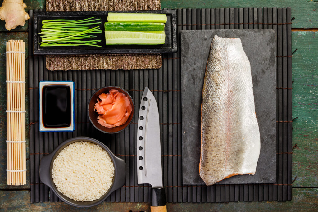 Traditional sushi ingredients: rice, salmon filet, ginger, cucumber, chives, soy sauce and bamboo mat for how to make sushi