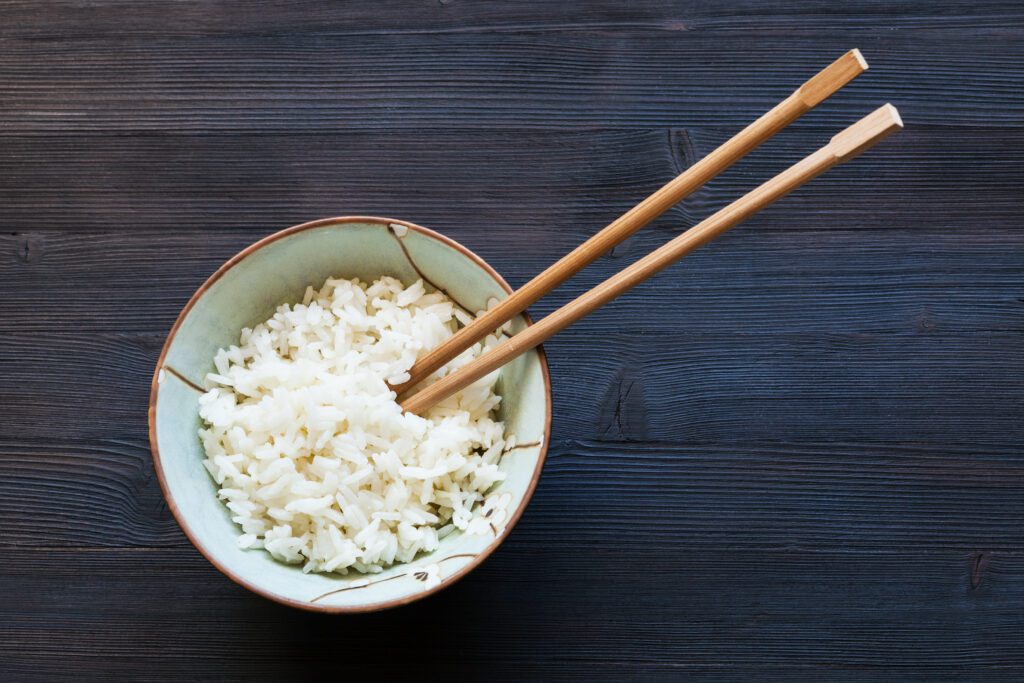 top view of chopsticks in bowl with boiled rice on dark wooden board